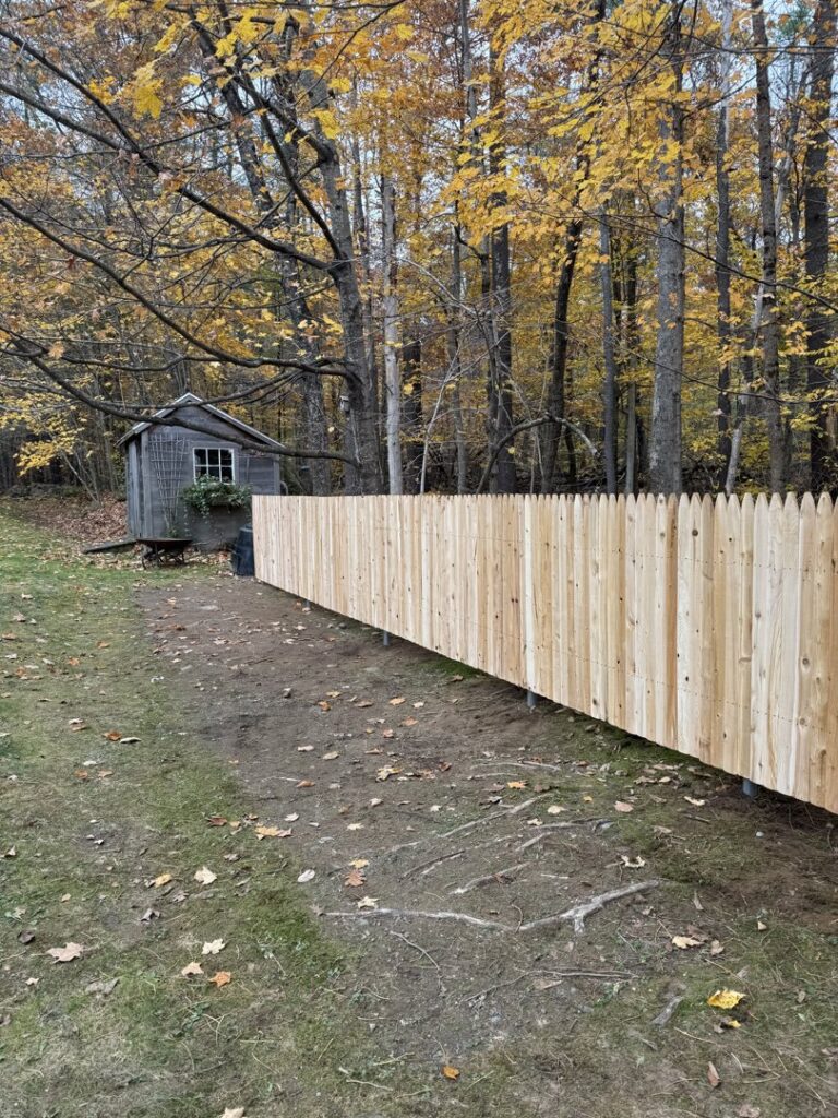 A newly installed wooden picket fence in a backyard with a shed and autumn trees by Triple P Fence in Augusta, ME.