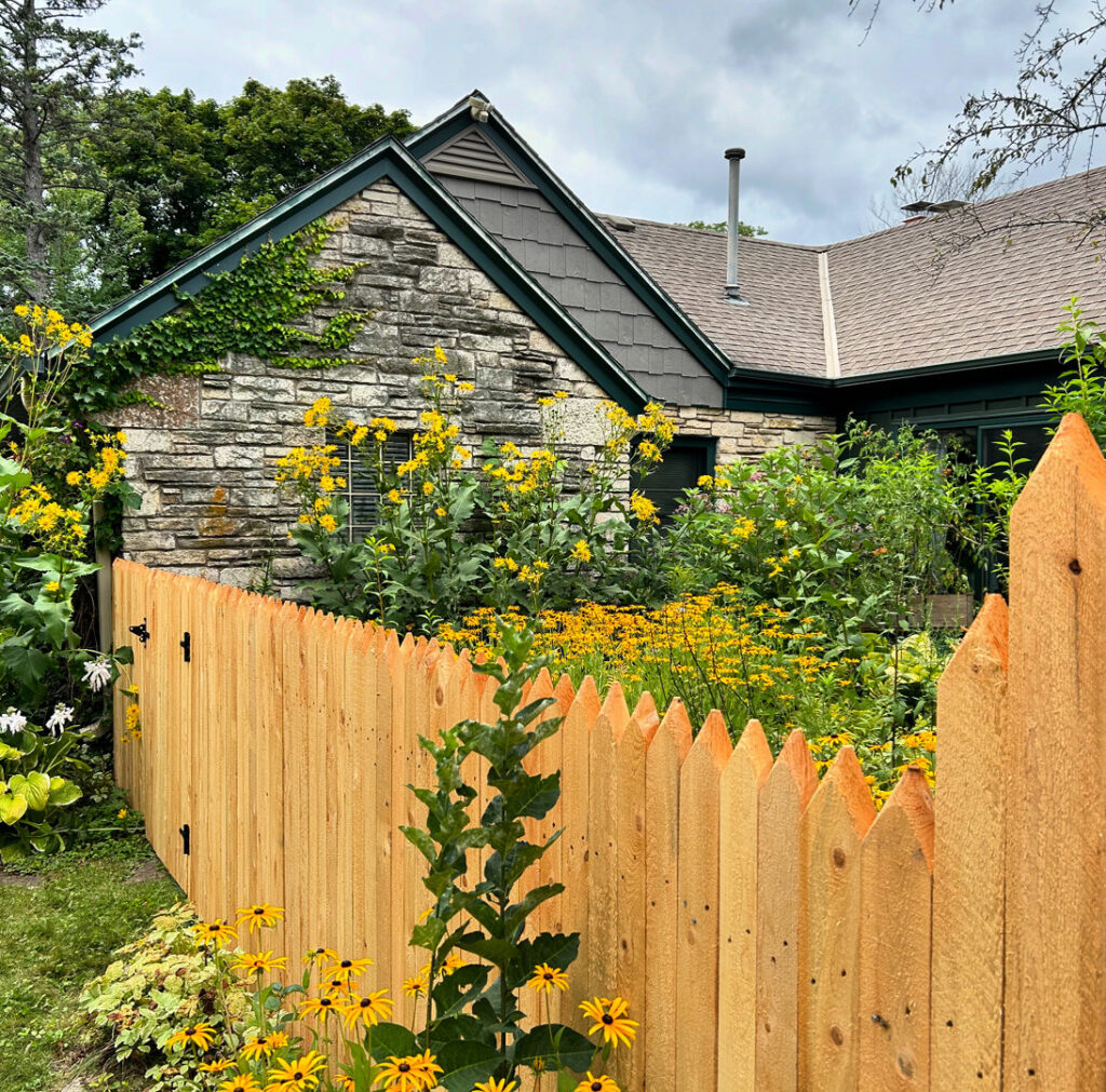 A charming wooden picket fence with pointed tops surrounding a vibrant garden by Chisel + Vine in Milwaukee, WI.