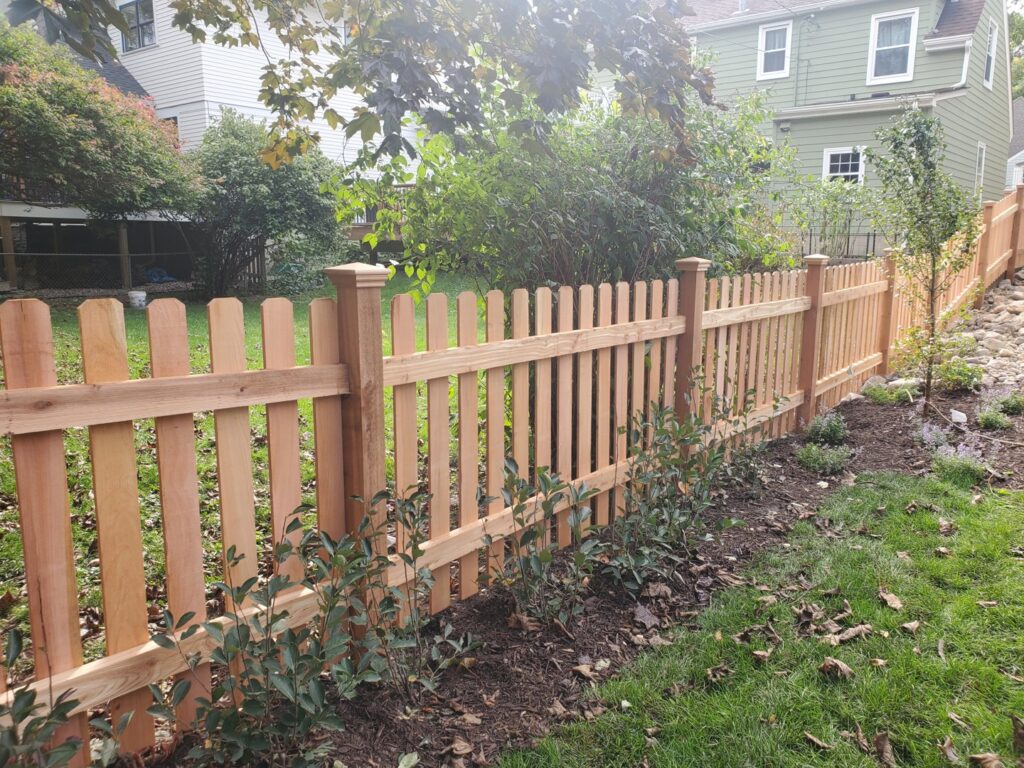 A charming wooden picket fence with decorative post caps bordering a garden by Top Notch Fence in East Bethel, MN