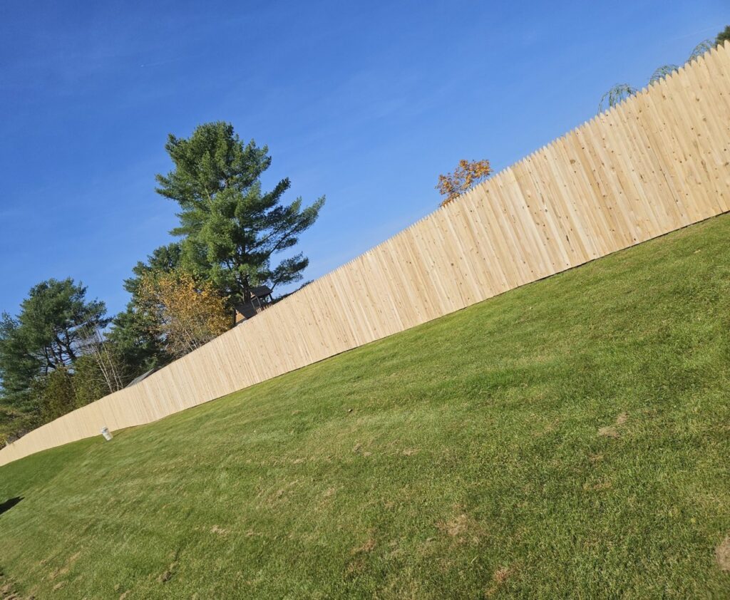 A long wooden picket fence installed on a sloped green lawn under a clear blue sky by Triple P Fence in Augusta, ME.