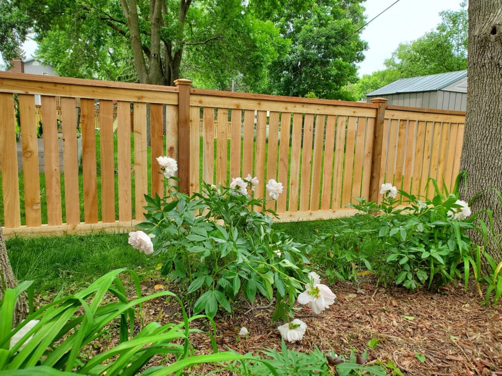 A beautiful wooden privacy fence with a decorative top rail and post caps, complemented by blooming flowers, by Top Notch Fence in East Bethel, MN.