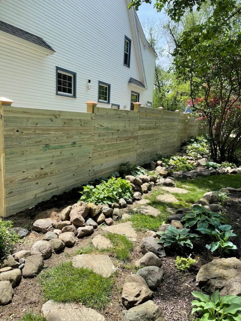 A wooden privacy fence with decorative post caps, bordering a landscaped garden, installed by NBBJ Fence LLC in Bluffton, IN.