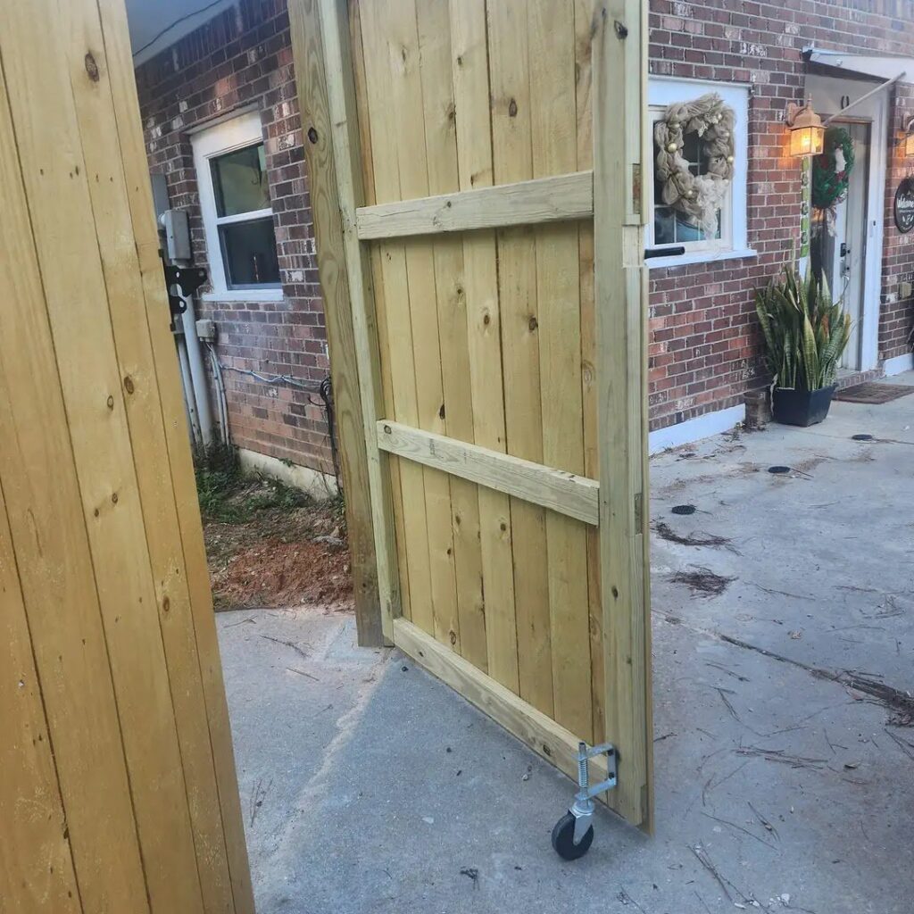 A newly installed wooden rolling gate with a wheel for easy access, next to a brick house, by Riggins Renovations LLC in Biloxi, MS.