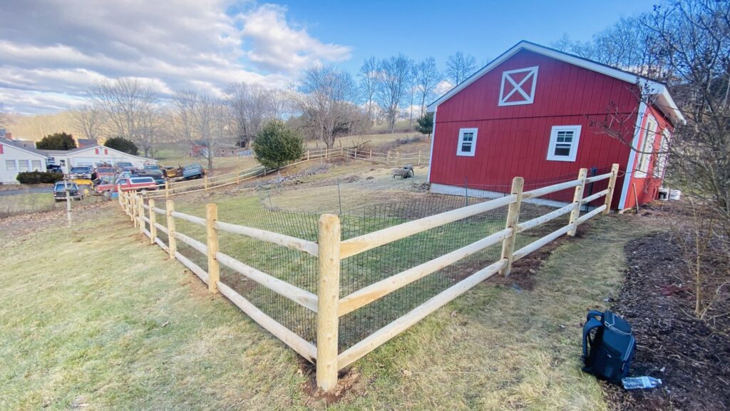 A wooden split rail fence with mesh installed near a red barn by White Eagle Fence Inc in New Haven, CT.