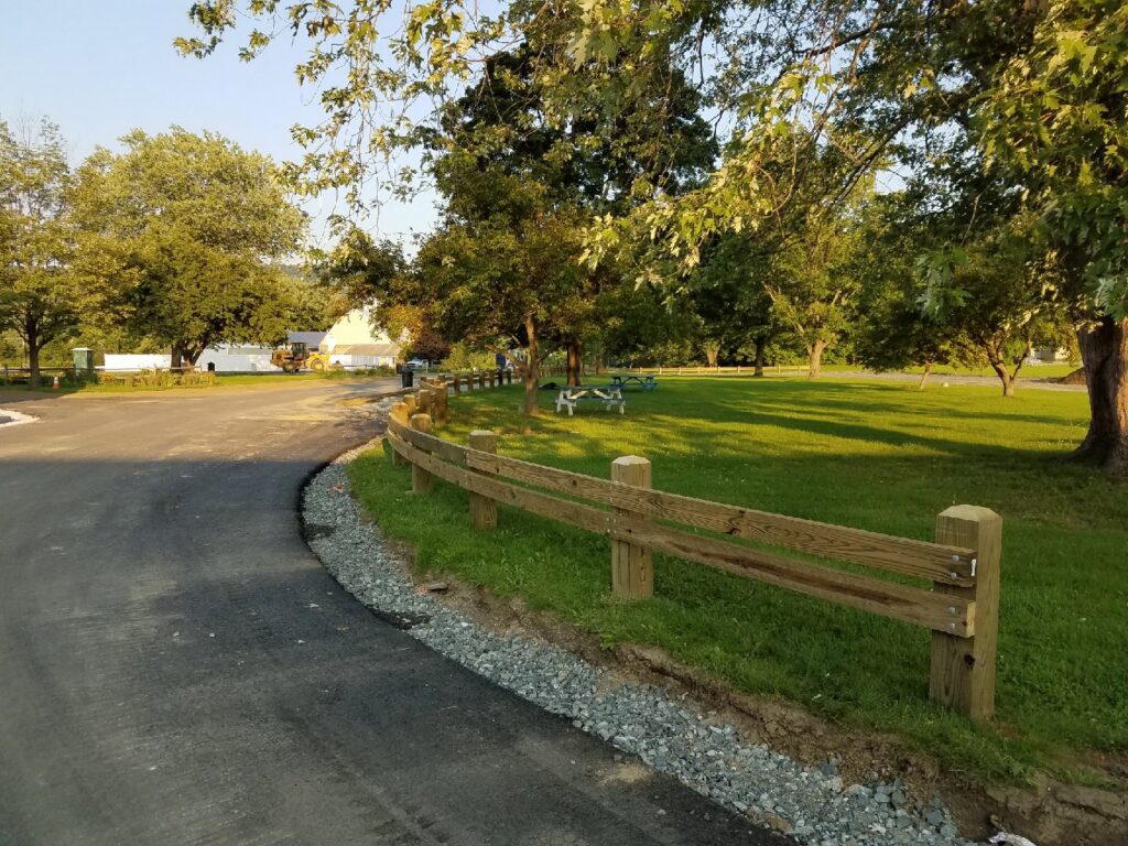A wooden split-rail fence installed along a paved path in a park by 802 Fence Company LLC in Shoreham, VT.