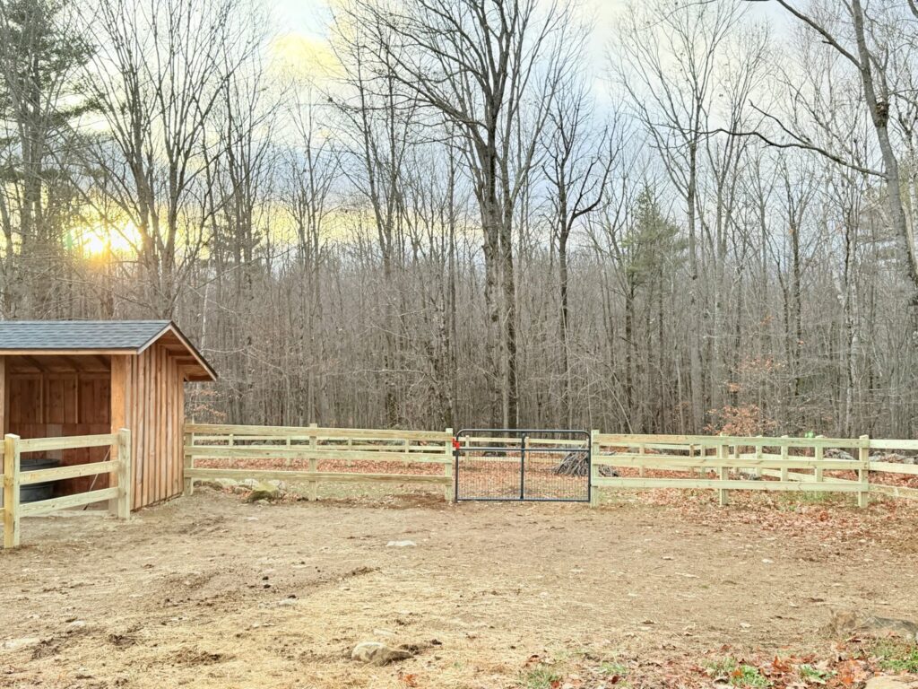A wooden split-rail fence with wire mesh and a metal gate in a rural setting by Ultra Fence LLC in Brookline, NH.