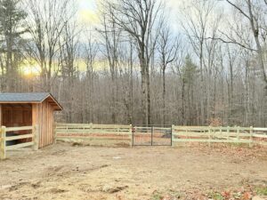 A wooden split-rail fence with wire mesh and a metal gate in a rural setting by Ultra Fence LLC in Brookline, NH.