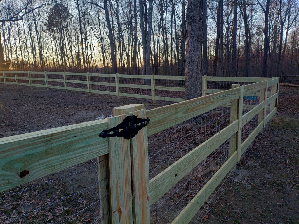 A newly installed wooden split-rail fence with wire mesh extending into a wooded area by Kelley Fence and Gate in Eva, AL.
