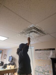 A worker on a ladder cleaning a ceiling air vent in an office, performed by Aspen Air Duct Cleaning in Methuen, MA.