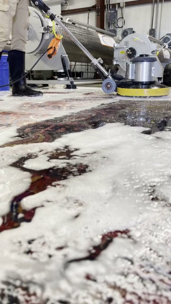 A worker using a floor buffer to clean a large area rug, with cleaning foam visible, at Advanced Cleaning Systems in Atlanta, GA.