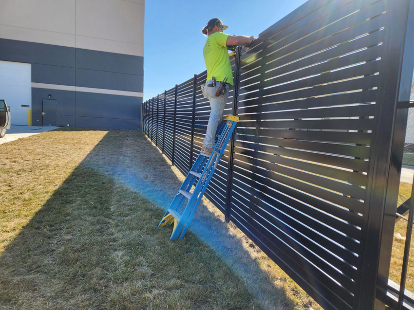 A worker on a ladder installing a black slatted fence for Van's Fence in Kansas City, MO.