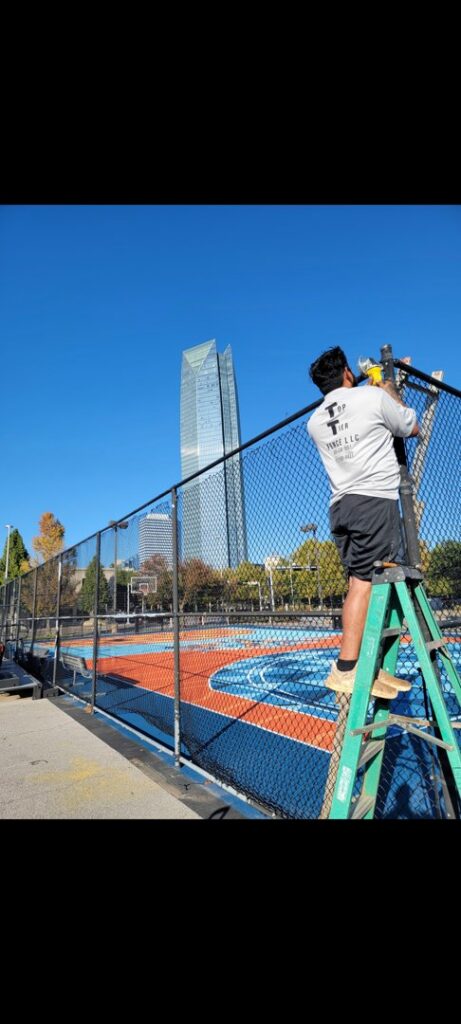 A worker on a ladder installing a chain link fence with the Oklahoma City skyline in the background by TOP TIER FENCE LLC.