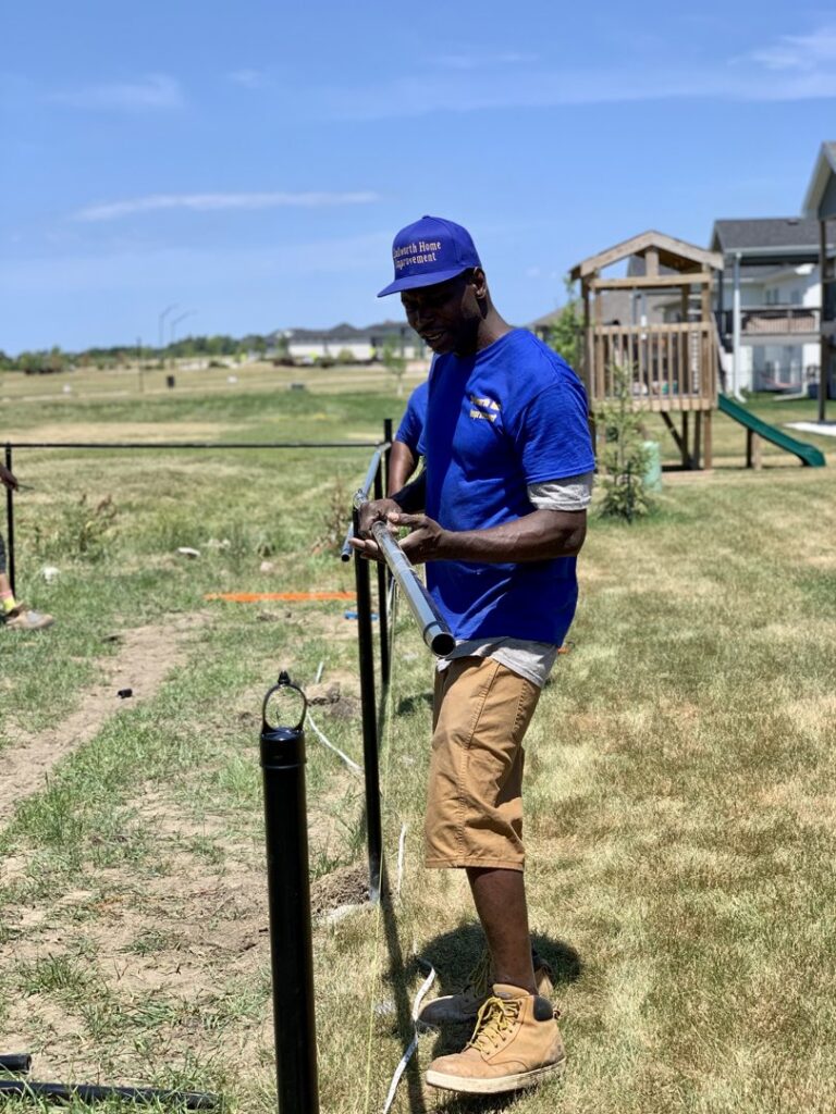 A worker from Stallworth Home Improvement installing a fence post in Des Moines, IA, under a clear blue sky.