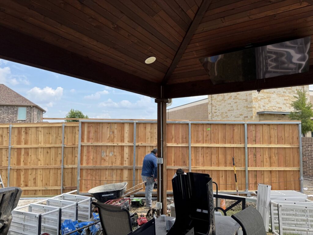 A worker installing wood panels onto metal posts for a privacy fence, viewed from under a patio, by Dallas Fence in Dallas, TX.