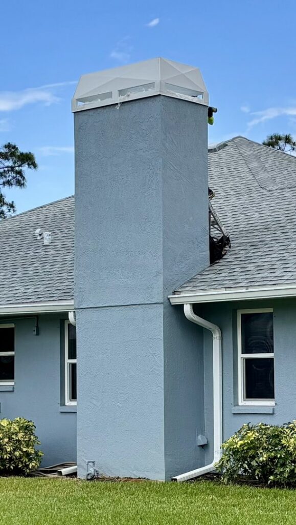 A worker on a ladder performing service on a tall blue chimney with a white cap for SWFL Chimney Caps Inc. in Fort Myers, FL.