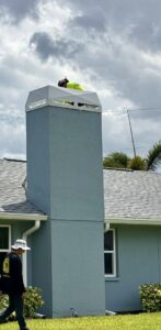 A worker on a ladder performing service on a tall chimney with a white cap for SWFL Chimney Caps Inc. in Fort Myers, FL.