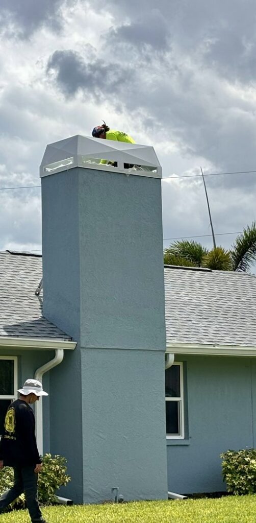 A worker on a ladder performing service on a tall chimney with a white cap for SWFL Chimney Caps Inc. in Fort Myers, FL.