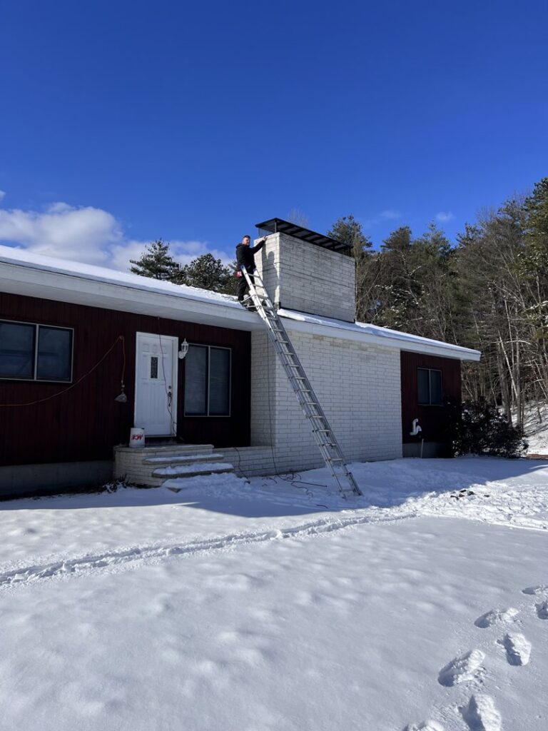 A worker on a ladder performing chimney service on a snowy roof for Granite State Chimney in Manchester, NH