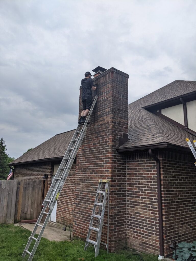 A worker on a ladder performing chimney service at the top of a brick chimney by Clean Sweep Chimney Service in Saint Charles, MO.