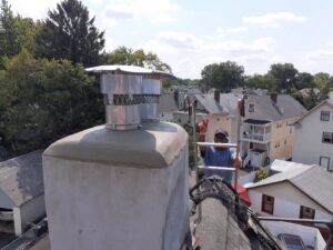 A worker on a ladder on a roof installing new chimney caps, performing a chimney service by Colombus Chimney in Elizabeth, NJ.
