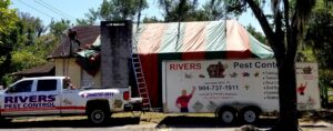 A worker on the roof of a house covered in a fumigation tent, with a Rivers Pest Control truck and trailer nearby in Jacksonville, FL