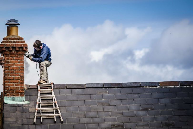 A worker on a ladder repairing a brick chimney on a roof for Pros Chimney Sweep Dryer Vent Cleaning in Chicago, IL.