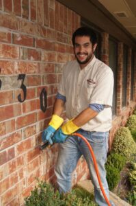 A smiling Reliable Termite of OKC technician spraying the foundation of a brick house in Oklahoma City, OK.