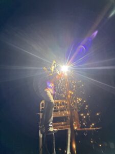 A worker welding a fence component at night, with bright sparks, for Cool Hand Fencing and Wyo War Wagons in Laramie, WY.