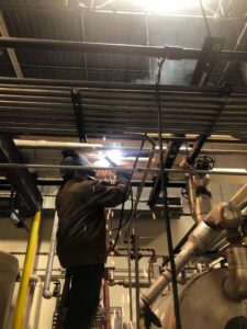 A worker welding pipes overhead in an industrial setting, part of an HVAC installation by R.J. O'Neil Inc. in Montgomery, IL.