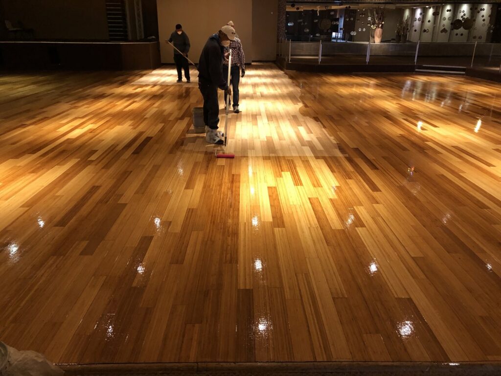 Workers applying a protective finish to a large wooden floor, a service provided by Carpets Texas in El Paso, TX.