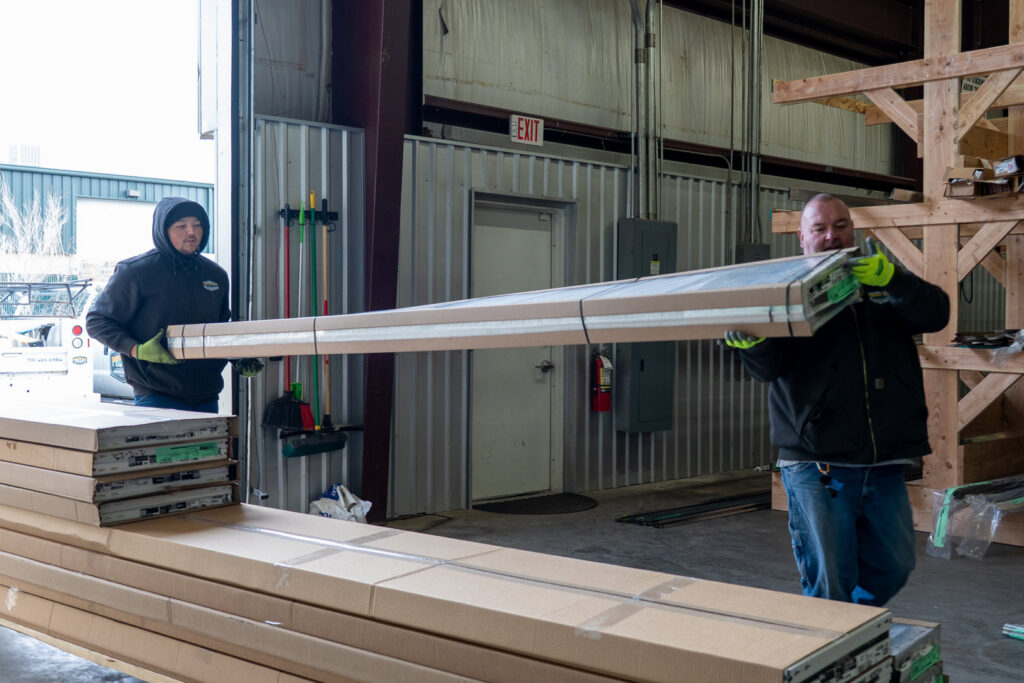 Two Prutch's Garage Door workers carrying a long, packaged garage door panel or track inside a warehouse in Colorado Springs, CO