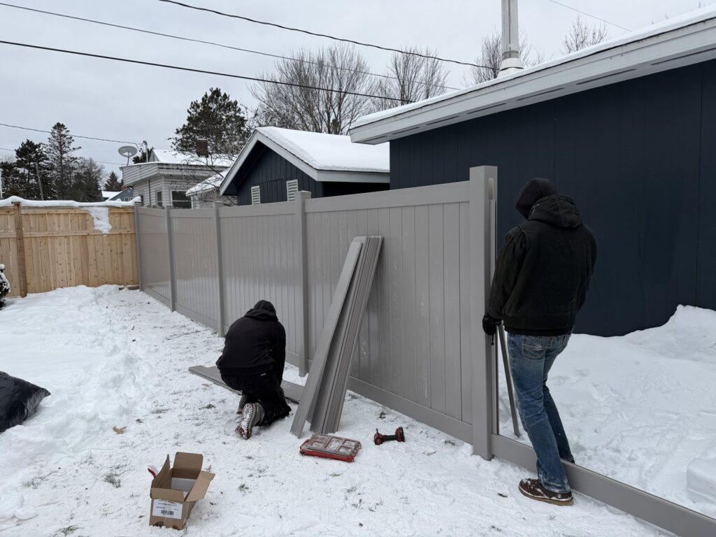 Workers installing a gray vinyl privacy fence in a snowy backyard by North Shore Fence Company in Centereach, NY.