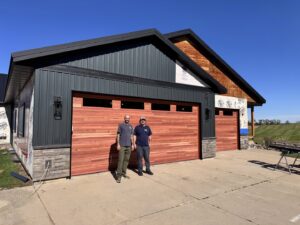 Two workers standing proudly in front of newly installed wood-look garage doors on a house under construction, by Madison Overhead Garage Door Services in Madison, WI.