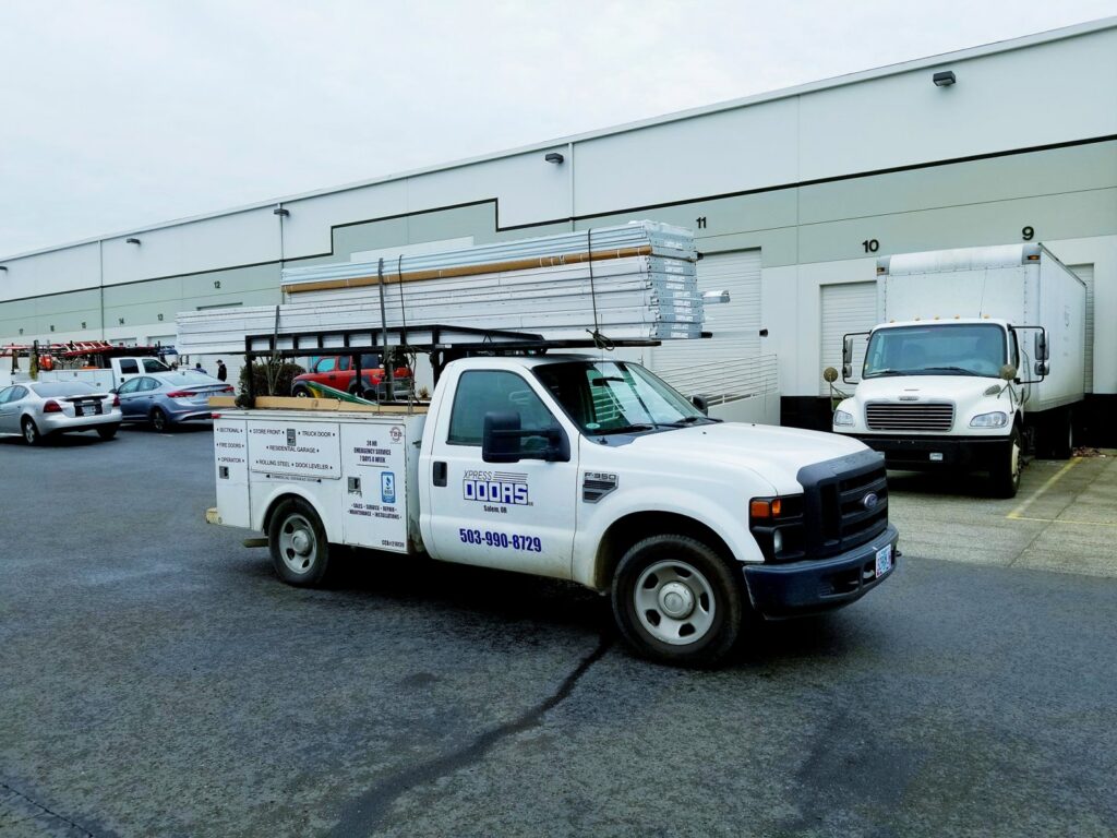 An Xpress Doors LLC service truck loaded with garage door panels, parked at a commercial job site in Salem, OR