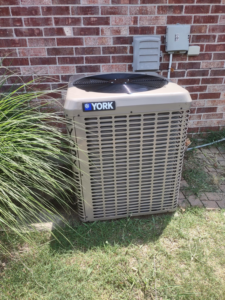 A new York brand outdoor AC condenser unit installed next to a brick house by Pettry Plumbing Heating & Air Inc in Bentonville, AR.