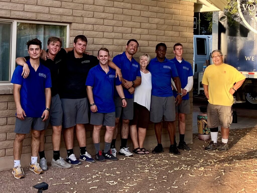 The Academy Movers team in blue shirts posing with two happy customers outside a home next to their moving truck in Gilbert, AZ.
