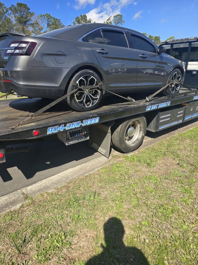 Adkison Towing flatbed truck transporting a grey sedan on a sunny day in Jacksonville, FL.