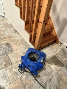 An air mover drying a tiled floor near a wooden staircase during water damage restoration by Restoration Alliance Services in Elk River, MN.