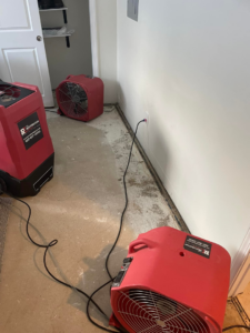 Red air movers and fans drying a concrete floor with visible water damage along the base of the wall, handled by The Restoration Experts in Waupaca, WI.