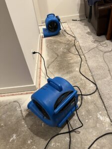 Two industrial air movers drying a concrete floor after carpet removal due to water damage, by Cowboy Certified Plumbing & Water Cleanup in Lee's Summit, MO.