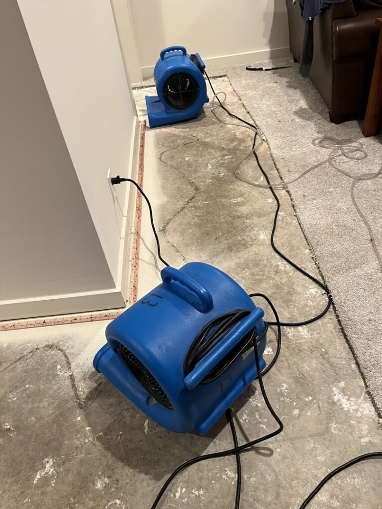 Two industrial air movers drying a concrete floor after carpet removal due to water damage, by Cowboy Certified Plumbing & Water Cleanup in Lee's Summit, MO.