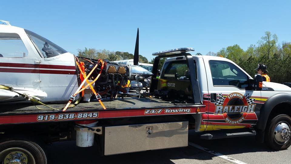 A tow truck from Raleigh Towing & Recovery transporting a small airplane with its engine exposed in Raleigh, NC.