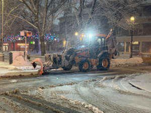 A backhoe actively plowing snow on a street at night for the City of Cambridge Department of Public Works in Cambridge, MA.