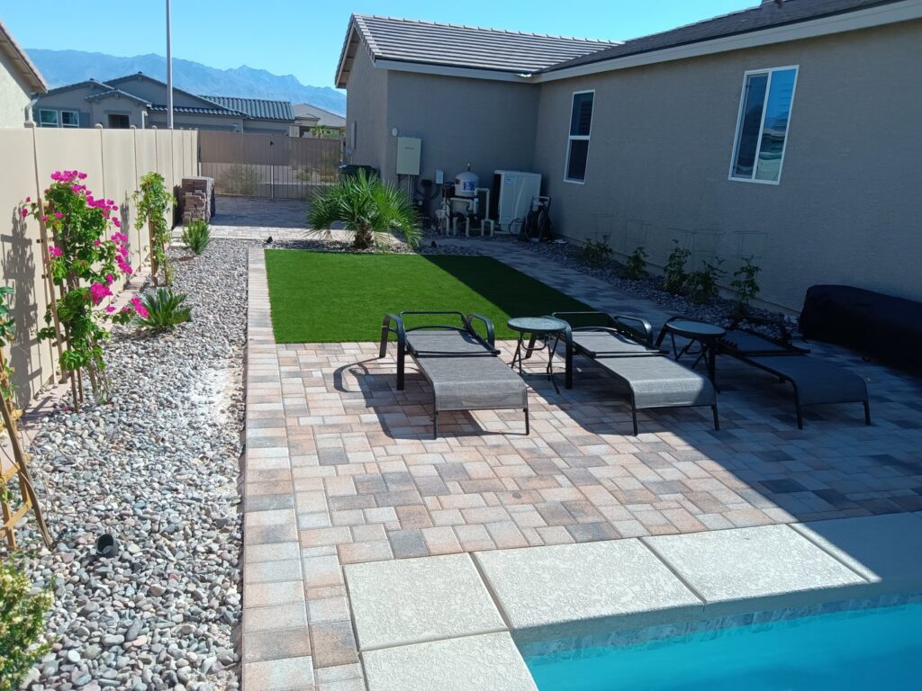 A backyard patio with pavers, artificial turf, lounge chairs, and a glimpse of a pool by Greencare.net POOL Builder in Las Vegas, NV