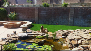 A backyard featuring a decorative pond, fire pit, and artificial grass installed by SYNLawn of New Mexico in Albuquerque, NM.