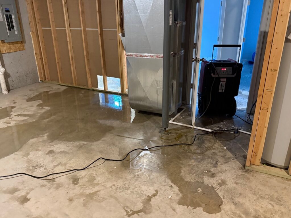 Standing water on a concrete basement floor with a dehumidifier running, showing water damage cleanup by Cowboy Certified Plumbing & Water Cleanup in Lee's Summit, MO.