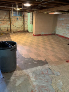 A basement floor showing extensive water damage with peeling tiles and exposed subfloor, undergoing remediation by Local Clean Up Specialists in Green Bay, WI.