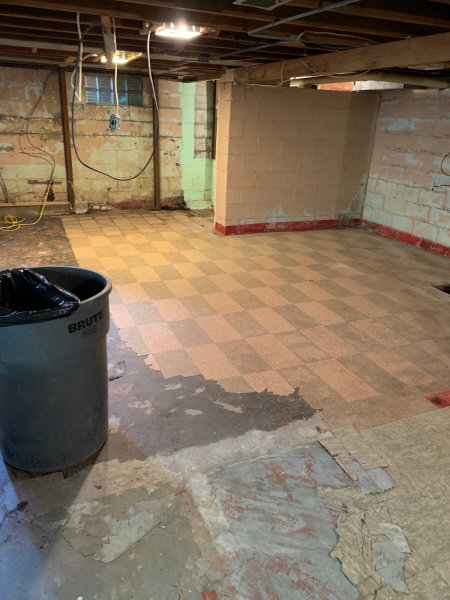 A basement floor showing extensive water damage with peeling tiles and exposed subfloor, undergoing remediation by Local Clean Up Specialists in Green Bay, WI.