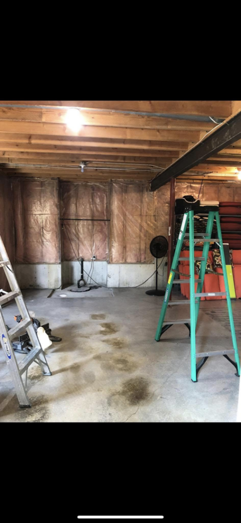 A basement undergoing water damage remediation with visible floor stains and a drying fan by Tules Remodeling LLC in Olathe, KS
