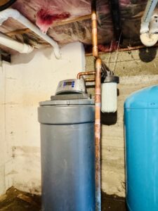 A water softener system and pressure tank in a basement with visible water stains on the floor and wall, indicating water damage, by The Plumbing Dr in Falls Church, VA